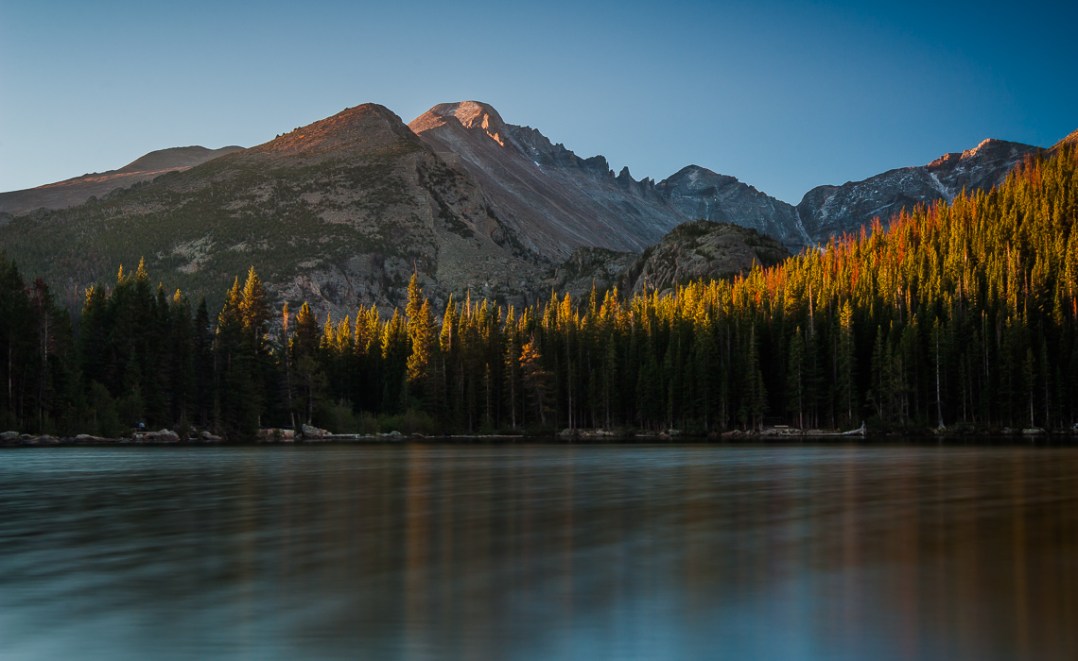 First light during early morning sunrise over Longs Peak, Rocky Mountain National Park. The peak and the forrest surrounding it are reflected as a rainbow spectrum in the throng if light ripples on the surface of Bear Lake.