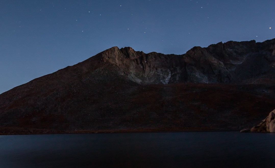 Mount Evans lit from the right by the final rays of sunlight during Astronomical sunset, and lit by the rising moon from the left. A few stars glitter overhead.